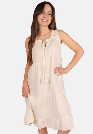 Femme portant une robe midi crème sans manches avec un col volanté et un lien à pompons, souriante avec une main sur la hanche sur fond blanc.