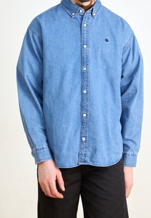 Mężczyzna ubrany w jasnoniebieską dżinsową koszulę zapinaną na guziki z długimi rękawami i czarne spodnie na jednolitym tle.