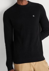 Svart ribbad stickad tröja med rundad halsringning och liten logotyp på bröstet. Långa ärmar och figursydd nederkant. Kombinerad med beige byxor.