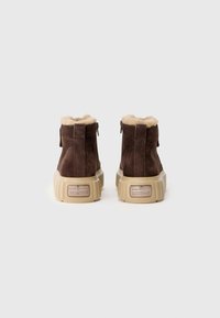Bottines en daim marron avec doublure en fausse fourrure crème, semelle en caoutchouc beige et une petite plaque logo à l'arrière. Détail zip à l'arrière.