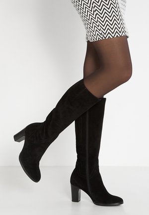Legs in sheer black tights wearing knee-high black suede boots with block heels and a white skirt with a black zigzag pattern.