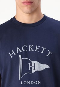 Camiseta de algodón azul marino con un gran diseño impreso de una bandera con el texto "H" y "HACKETT LONDON" en blanco. Manga corta y cuello redondo.