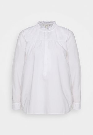 Blouse blanche à manches longues avec un col montant à volants, des plis verticaux sur le haut de la poitrine et une patte de boutonnage cachée à l’avant.