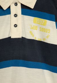 Stripet polo skjorte i marineblå, blå og kremfarge. Har tre knapper og en trykt gul etikett med "1997" og "Gooods Dept."