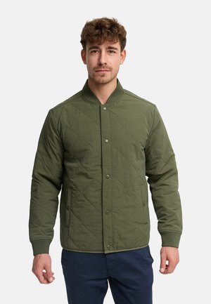 Mand iført en quiltet olivengrøn skjortejakke med knapper, ribbet kant ved ærmer og krave, stående mod en ensfarvet lys baggrund.
