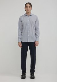 Un homme se tient face à l'avant, portant une chemise à manches longues à motifs légers, un pantalon de costume foncé et des chaussures noires à lacets, devant un fond uni.