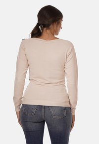 Maglione beige a maniche lunghe con scollatura rotonda, polsini e orlo a costine. Il tessuto è liscio con un design aderente. Indossato con jeans blu.
