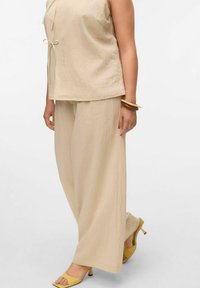 Haut sans manches beige texturé avec un détail de nœud à l'avant, associé à un pantalon large beige décontracté. Porté avec des escarpins jaunes à bout découvert.