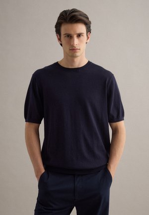 Joven con cabello oscuro que lleva un suéter azul marino de manga corta y pantalones azul marino, de pie con las manos en los bolsillos sobre un fondo liso.