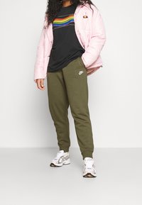 Vrouw in een roze pufferjack, zwart shirt met regenboogstrepen, olijfgroene Nike-sweatpants en witte sneakers, staande tegen een witte achtergrond.