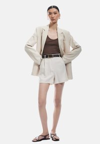 Cremefarvet blazer over en brun ribbet tanktop, bælteholdte hvide shorts og sorte strappy sandaler. Glat stof og tætsiddende pasform med minimale accenter.