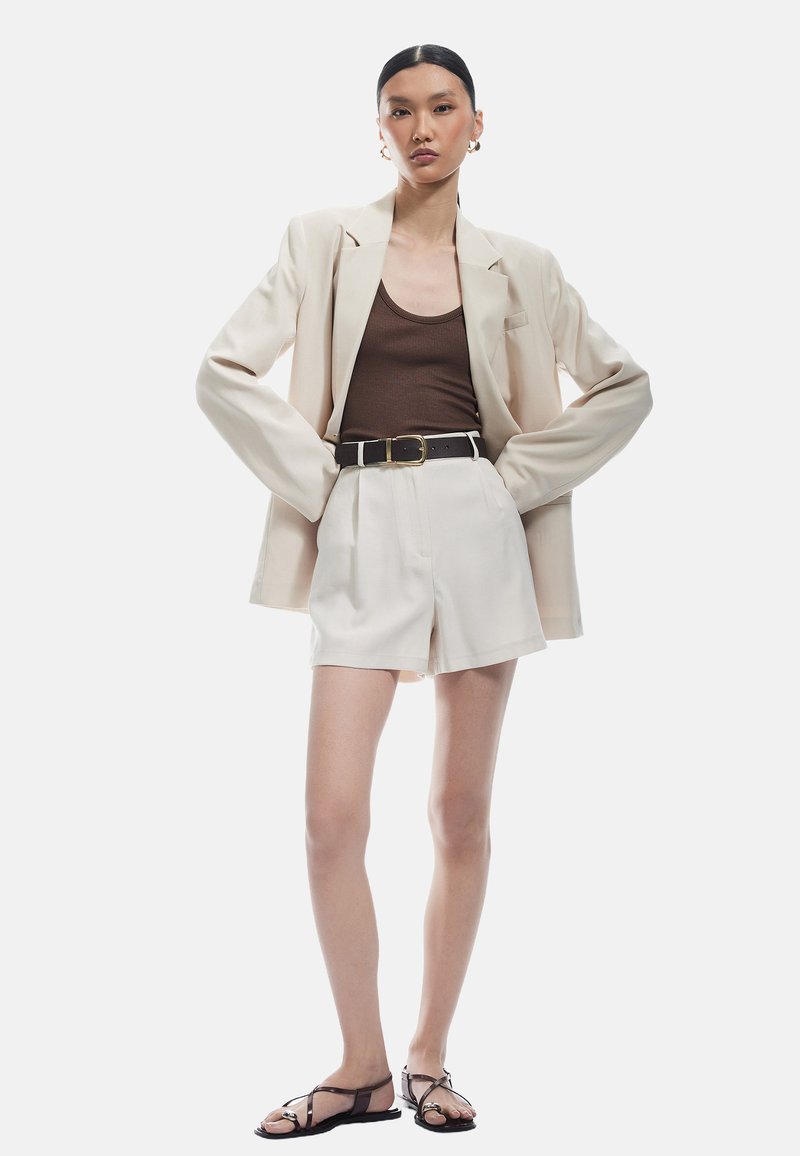 Cremfarbener Blazer über einem braunen gerippten Oberteil, geschnürte weiße Shorts und schwarze Riemchensandalen. Glatter Stoff und taillierte Passform mit minimalen Akzenten.