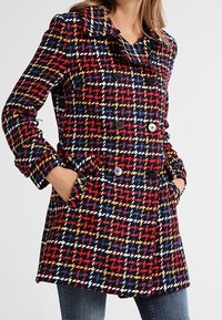 Dvojřadý kabát v pestrobarevném vzoru houndstooth v červené, námořnické modré, bílé a žluté barvě. Má velké černé knoflíky a boční kapsy.