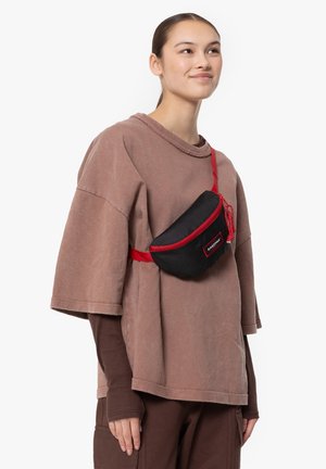 Zwart stoffen heuptasje met rode rits en band, met een gebogen vorm en logo-patch, gedragen over een losse, taupe oversized t-shirt.