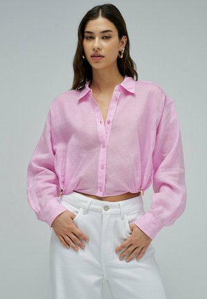Femme portant une chemise ample rose pâle boutonnée, rentrée dans un pantalon blanc taille haute, debout avec les mains près des poches.
