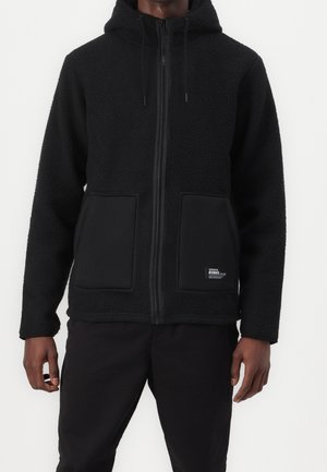 Man wearing black zip-up fleece hoodie with front pockets and hood, paired with black pants against a plain background.