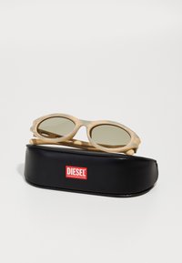 Ochelari de soare bej cu rame rotunde și lentile verzi, așezați pe o carcasă neagră cu un logo roșu "DIESEL".