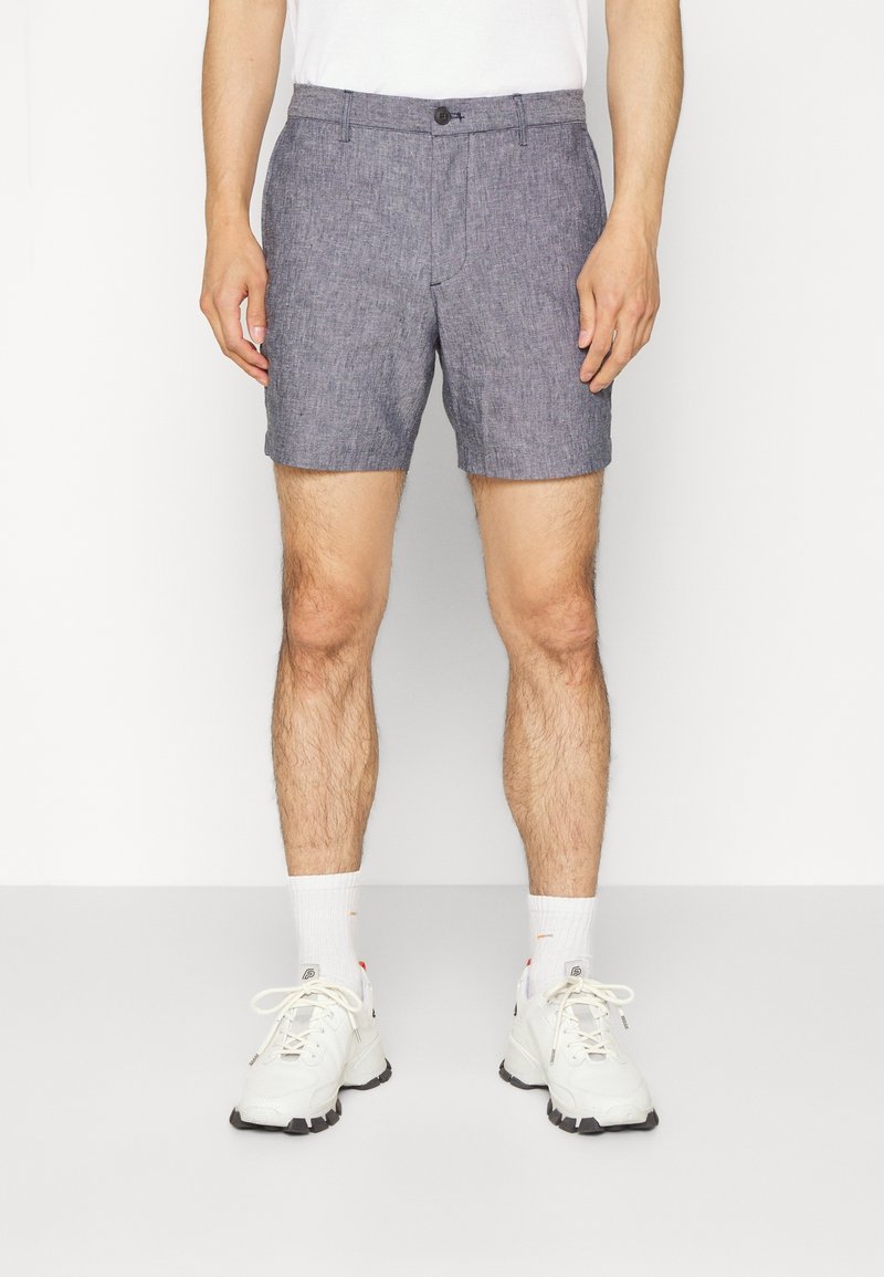 Pánské šedé pletené šortky s plochým předním dílem, zapínáním na knoflík a bočními kapsami. Nosené s bílými sportovními botami a ponožkami do poloviny lýtek.