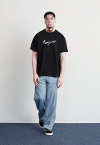 Zwarte katoenen t-shirt met witte scriptlogo, gecombineerd met lichtblauwe wijde broek en zwarte sneakers, staand tegen een effen achtergrond.
