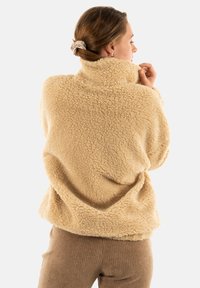 Pull beige in fleece texturizzato con colletto alto e fit ampio. Il tessuto ha un aspetto morbido, abbinato a pantaloni marroni a costine.