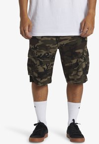 Cargo shorts in een groen en zwart camouflagepatroon van stof, met meerdere zijzakken en een losse pasvorm. Draag het met witte sokken en zwarte schoenen.