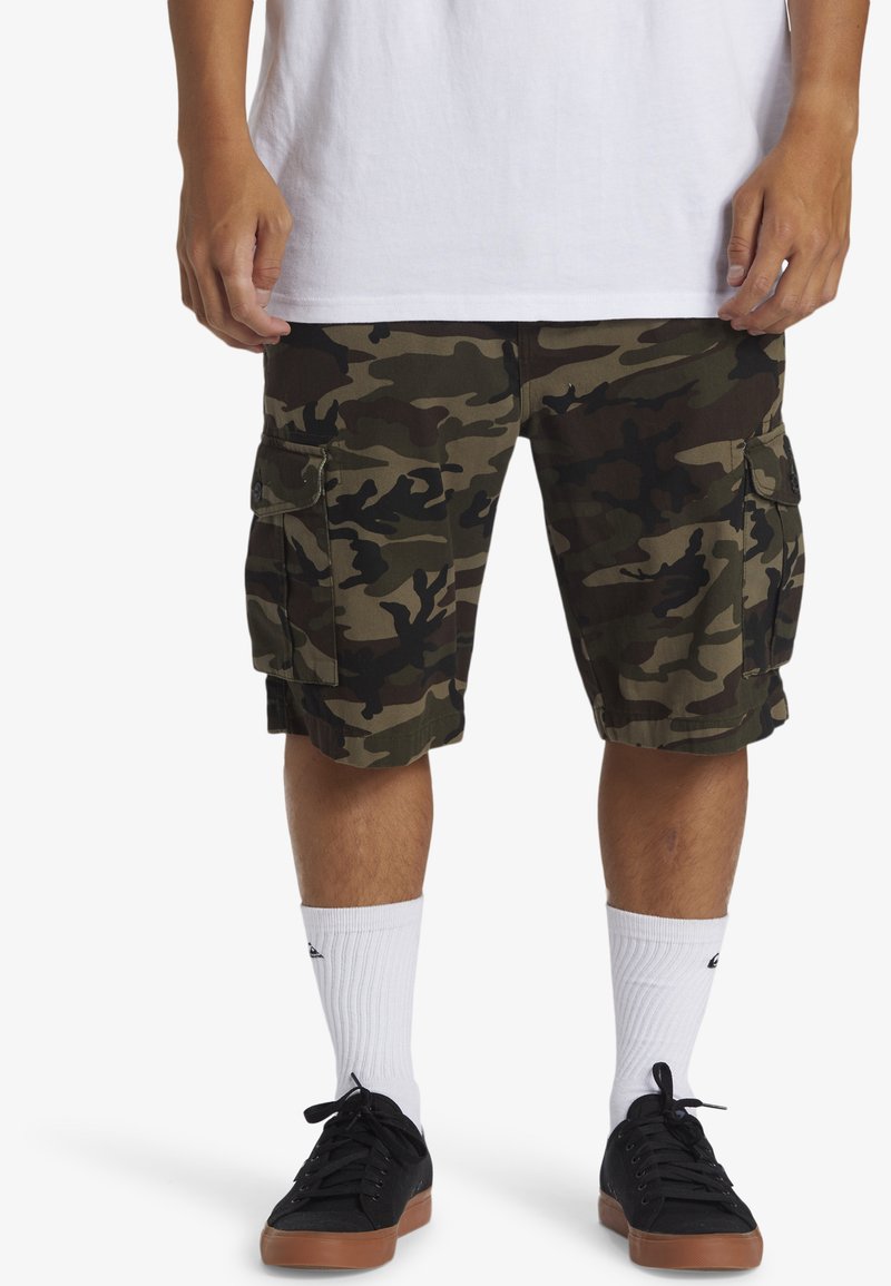 Cargo shorts in een groen en zwart camouflagepatroon van stof, met meerdere zijzakken en een losse pasvorm. Draag het met witte sokken en zwarte schoenen.