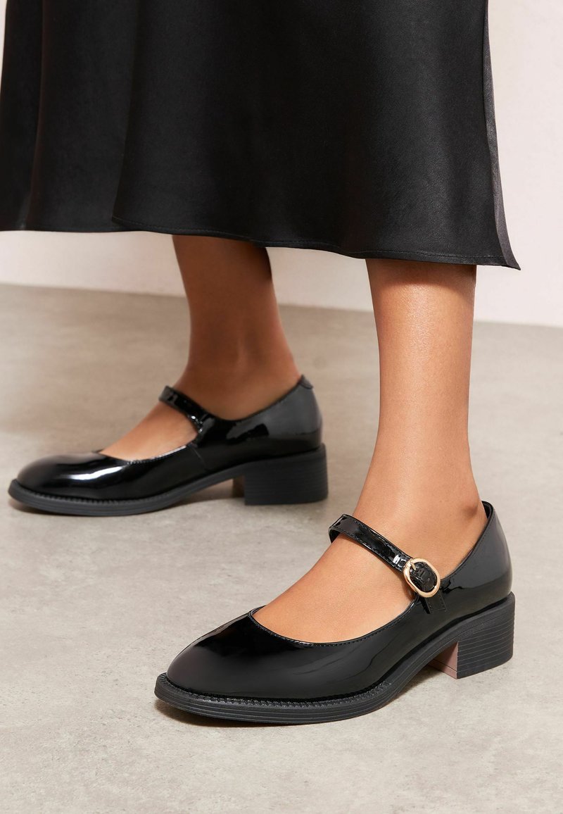 Črni lakirani Mary Jane čevlji z okroglo konico, nizkim blok peto, trakom okoli gležnja in okroglim zaponkom. Gladek videz in sijaj.