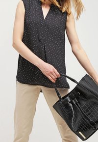 Blusa nera senza maniche con pois bianchi, dotata di scollatura a V. Pantaloni beige e borsa nera texturizzata con chiusura a coulisse.