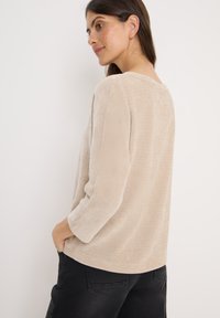 Haut en tricot texturé beige avec une coupe ample, des manches 3/4 et un col large, présenté sur un modèle de profil devant un fond uni.