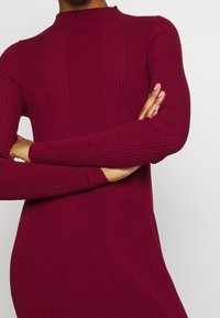 Robe en maille côtelée bordeaux avec manches longues, col haut et textures verticales, mettant en valeur une coupe ajustée et un tissu lisse.