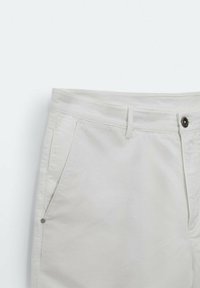 Pantalon en coton de couleur claire avec une partie avant plate, deux poches latérales et une fermeture à bouton noir. Texture lisse et design ajusté.