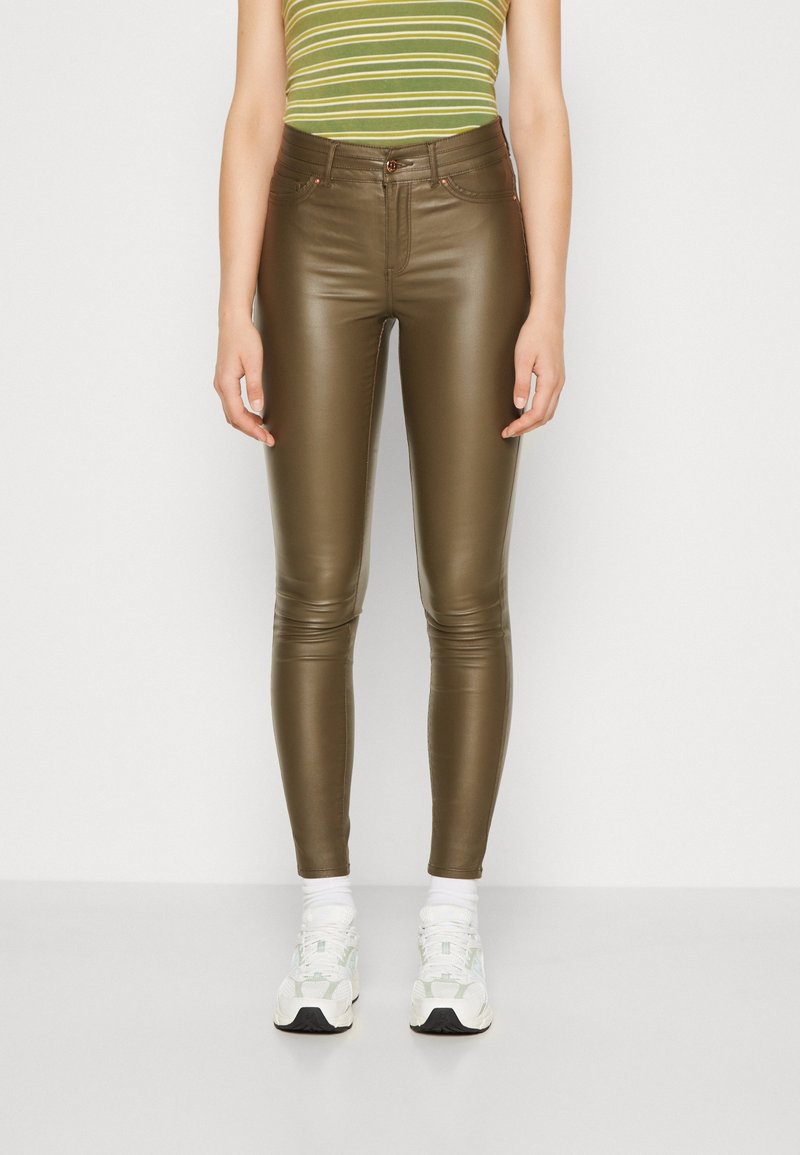Femme portant un pantalon skinny en cuir vert olive, des baskets blanches et un haut sans manches rayé vert, debout devant un fond clair uni.