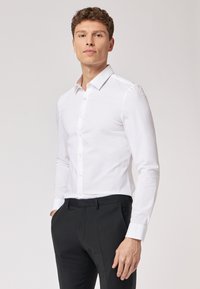 Camicia bianca a maniche lunghe con colletto classico, chiusura frontale con bottoni e vestibilità slim. Il tessuto ha una texture liscia e leggermente lucida.