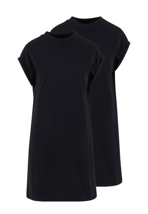 Femme aux cheveux bouclés portant une robe noire à manches courtes, debout devant un fond gris clair uni.