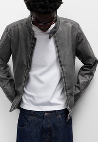 Uomo che indossa una giacca di pelle grigia sopra una t-shirt bianca e jeans blu, che guarda in basso, con le mani parzialmente nelle tasche della giacca, su uno sfondo bianco.