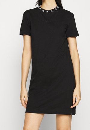 Kvinde iført en kort sort t-shirt-kjole med CK-logo mønster omkring halsudskæringen, stående med armene afslappet ned langs siden.