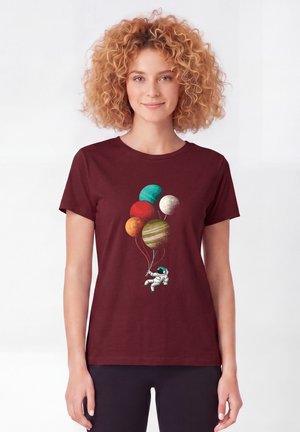 Femme aux cheveux bouclés portant un t-shirt bordeaux avec un astronaute tenant des ballons en forme de planète qui flottent vers le haut.