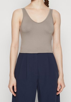 Femme portant un top court sans manches taupe à col en V et un pantalon large taille haute bleu marine, debout devant un fond uni.