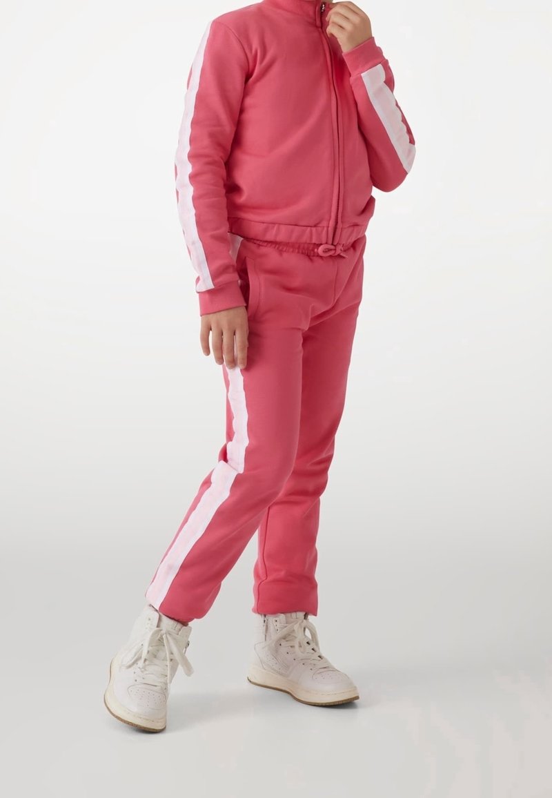 Enfant portant un survêtement rose avec des rayures latérales blanches et des baskets montantes blanches, debout et tenant la fermeture éclair de la veste près du visage.