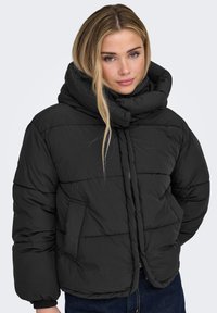 Musta toppatakki, jossa on korkea kaulus ja vetoketju, vaakasuora tikkaus ja sivutaskut. Valmistettu sileästä, toppatusta materiaalista.