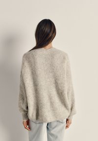 Svetlo sivi, prevelik pleteni pulover s teksturiranim vzorcem, padajočimi ramenmi in rebrastimi manšetami. Nošen z svetlo modrimi kavbojkami.