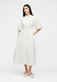 Γυναίκα που φοράει λευκό μίντι φόρεμα με φουσκωτά μανίκια, μυτερό γιακά, κυματιστή ραφή και λευκά χαμηλά τακούνια, στέκεται με τα χέρια στις τσέπες.