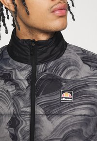 Svart och grå mönstrad puffjacka med hög krage, med en dragkedja fram och en Ellessee-logotyp på bröstet.