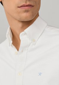 Nærbillede af en mands underansigt og hals iført en hvid skjorte med knapper og et lille lyseblåt broderet logo på brystet.