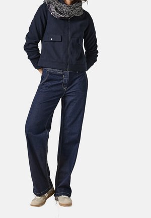 Person trägt eine dunkelblaue Jacke, einen gemusterten Schal, weit geschnittene dunkle Jeans und helle beige Sneaker, steht mit den Händen in den Taschen.