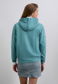 Woman facing away wearing teal hoodie over white shirt with gray zebra print skirt standing against plain light background.