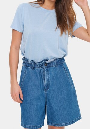 Vrouw draagt een lichtblauw T-shirt met korte mouwen, gestopt in een hooggesneden, loszittende blauwe denimshort met een gerimpelde tailleband.
