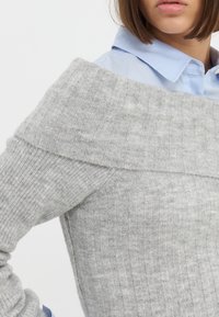 Maglione grigio a coste con design off-shoulder. Tessuto morbido, maniche aderenti e scollatura ampia. Indossato sopra una camicia colletto azzurra.