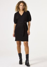 Vestido negro de manga corta con escote en forma de V, cintura ajustada con lazo y dobladillo a la altura de la rodilla. Combinado con botas negras hasta el tobillo. Acabado de tela suave.