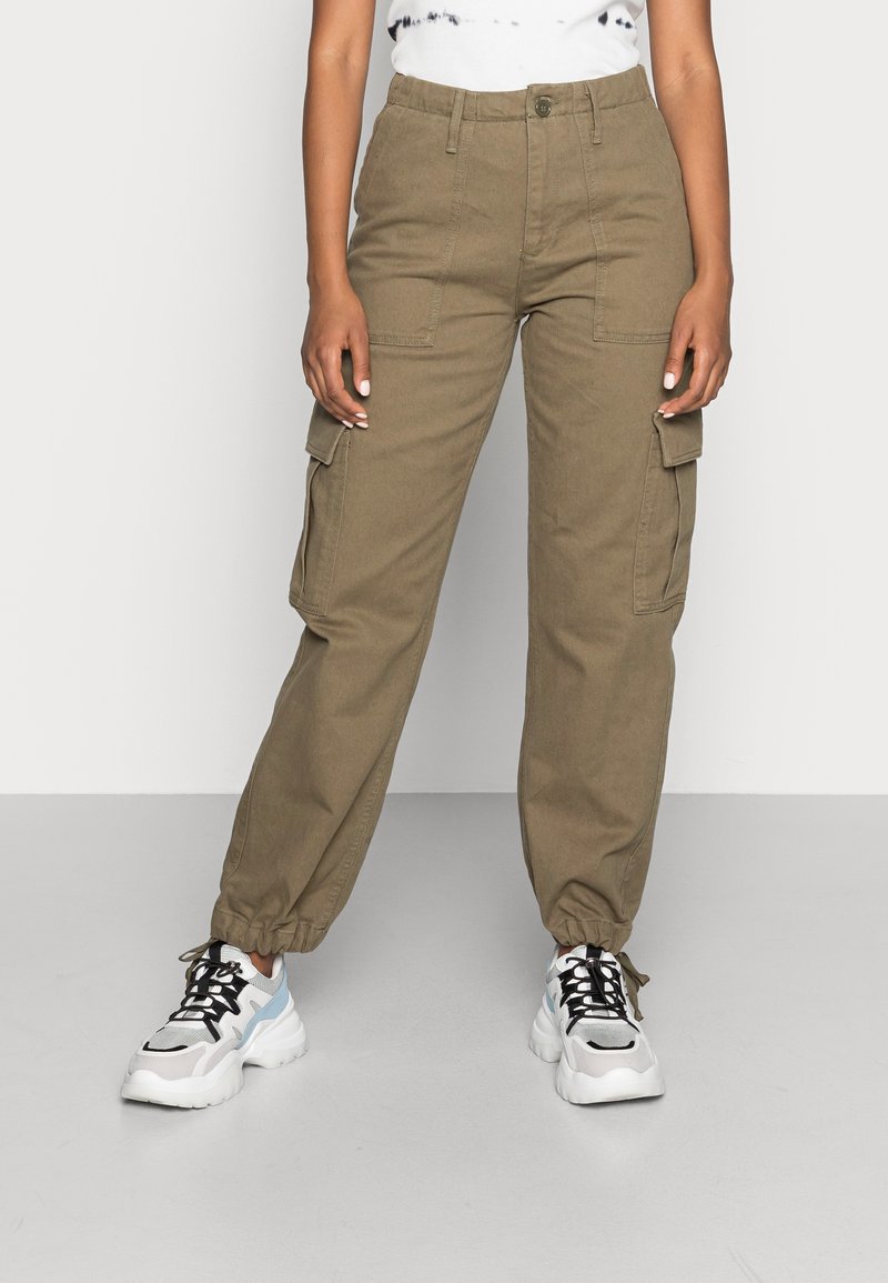 Personne portant un pantalon cargo kaki avec des poignets élastiques et des baskets blanches avec des lacets noirs, debout devant un fond uni.